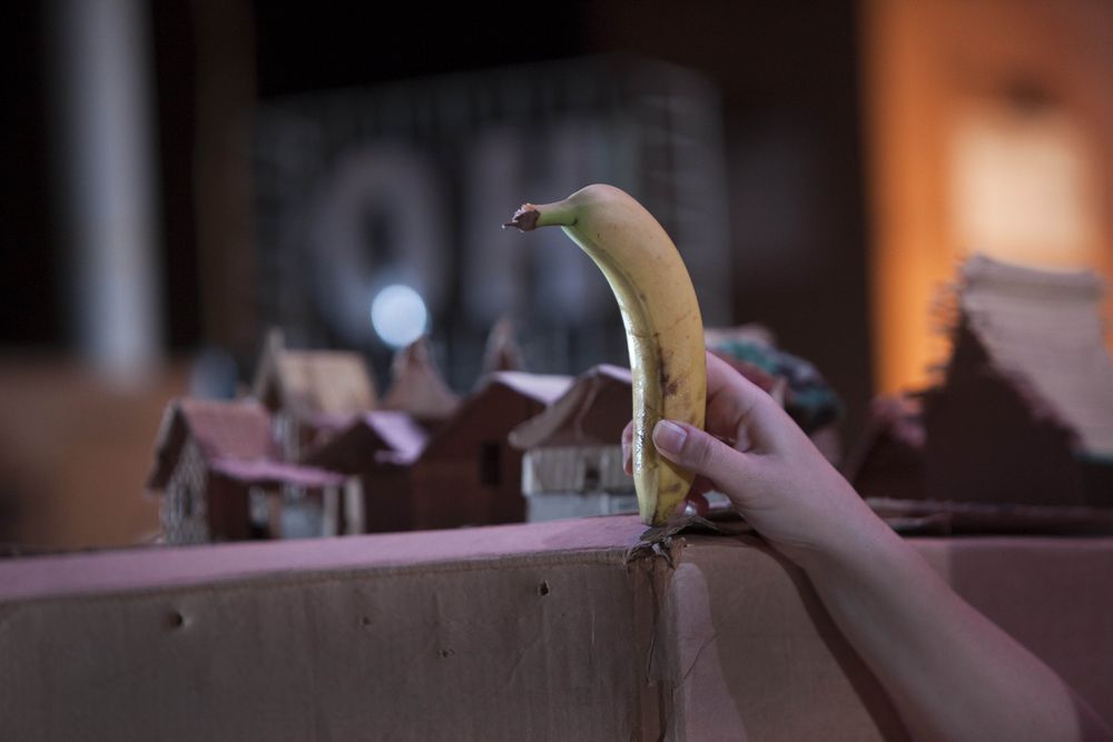 Een hand houdt een banaan omhoog boven een dorpje van kartonnen huisjes, met op de achtergrond het lichtbord OH.