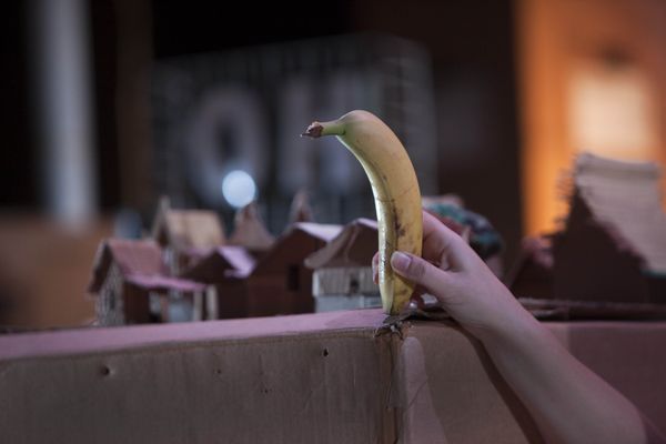 Een hand houdt een banaan omhoog boven een dorpje van kartonnen huisjes, met op de achtergrond het lichtbord OH.