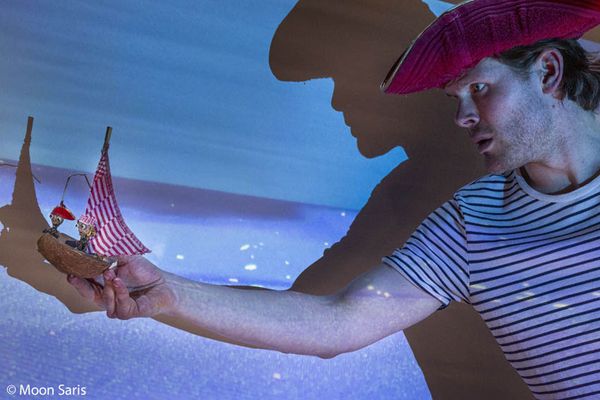 Een speler met een rode steek houdt een bootje van een kokosnoot met een gestreept zeil op zijn hand, tegen een blauw geprojecteerde zee.