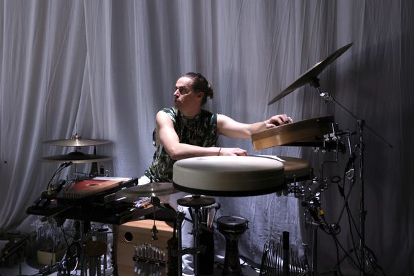 De muzikant speelt op zijn slagwerk van trommels, bekkens en belletjes voor een grijs gordijn.