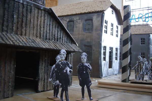 Twee uitgeknipte foto's van bewakers staan in de kampstraat tussen de barakken, rechts is het bord bij de poort te zien.