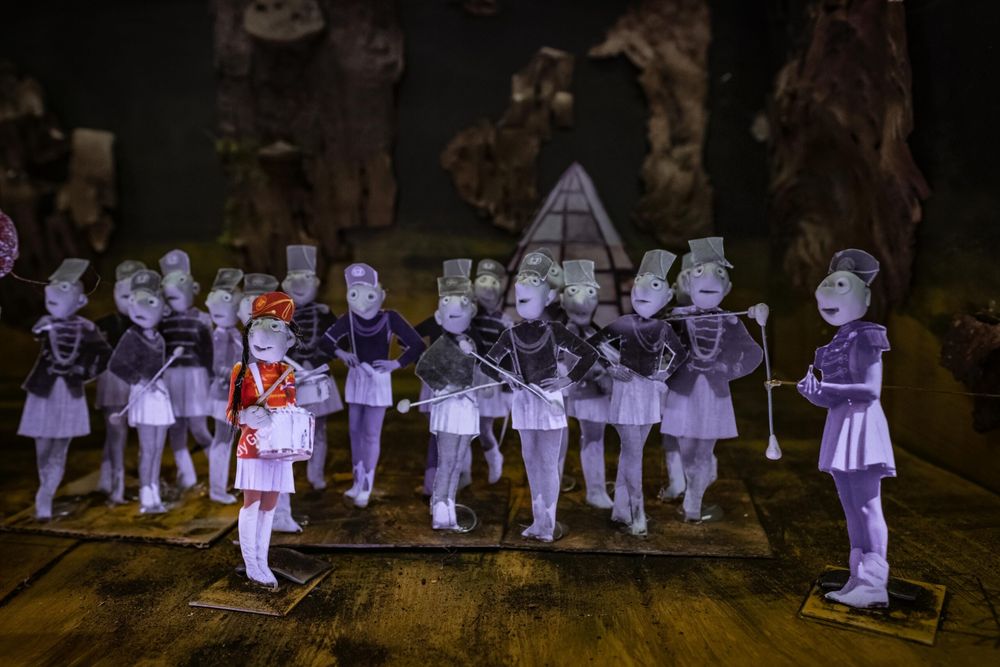 Uitgeknipte papieren figuurtjes vormen een showband op een houten vloer; alle majorettes zijn lila-grijs, één meisje in een rood uniform staat vooraan in kleur.