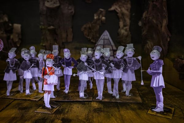 Uitgeknipte papieren figuurtjes vormen een showband op een houten vloer; alle majorettes zijn lila-grijs, één meisje in een rood uniform staat vooraan in kleur.