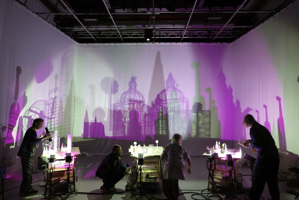 Wijd beeld van de studio: vier spelers werken aan lichttafels, op het grote doek een paarsgroene skyline van koepels en kooien.