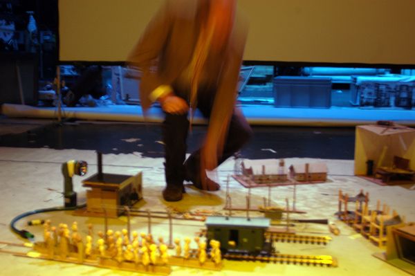 Een speler stapt over het miniatuurdecor met de treinwagon en de houten poppetjes, zijn gestalte vervaagd door de beweging.