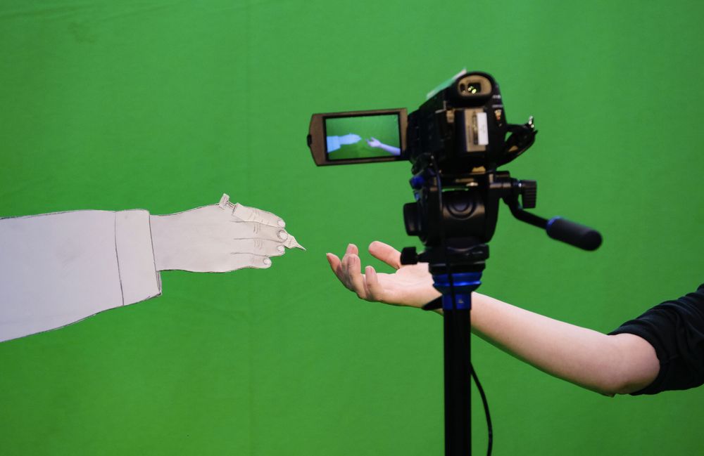 In de green screen-studio reikt een papieren hand naar een echte hand; ertussen staat de camera die het opneemt.