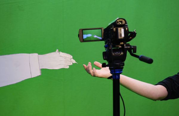 In de green screen-studio reikt een papieren hand naar een echte hand; ertussen staat de camera die het opneemt.