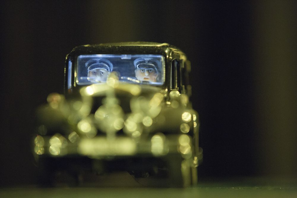 Een miniatuurauto komt met brandende koplampen recht op de camera af; achter de voorruit zitten twee poppetjes met helm en stofbril.