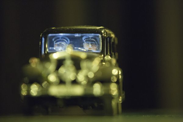Een miniatuurauto komt met brandende koplampen recht op de camera af; achter de voorruit zitten twee poppetjes met helm en stofbril.