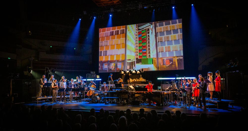 Wijd beeld van de concertzaal: het ensemble op het podium onder een scherm met een kleurige gevel van een flatgebouw.