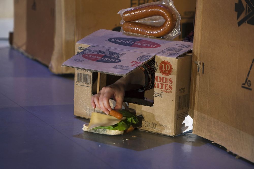 Een hand komt uit een gat in een kartonnen doos en grijpt naar een belegd broodje; boven op de doos ligt een rookworst.