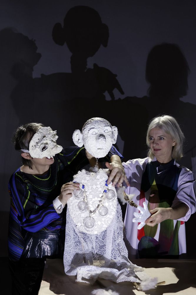 Twee speelsters houden een wit papieren beeld met een masker overeind; hun eigen schaduwen vallen groot op het doek erachter.