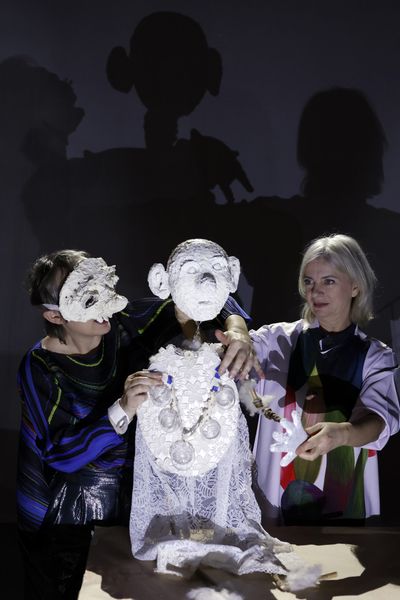 Twee speelsters houden een wit papieren beeld met een masker overeind; hun eigen schaduwen vallen groot op het doek erachter.