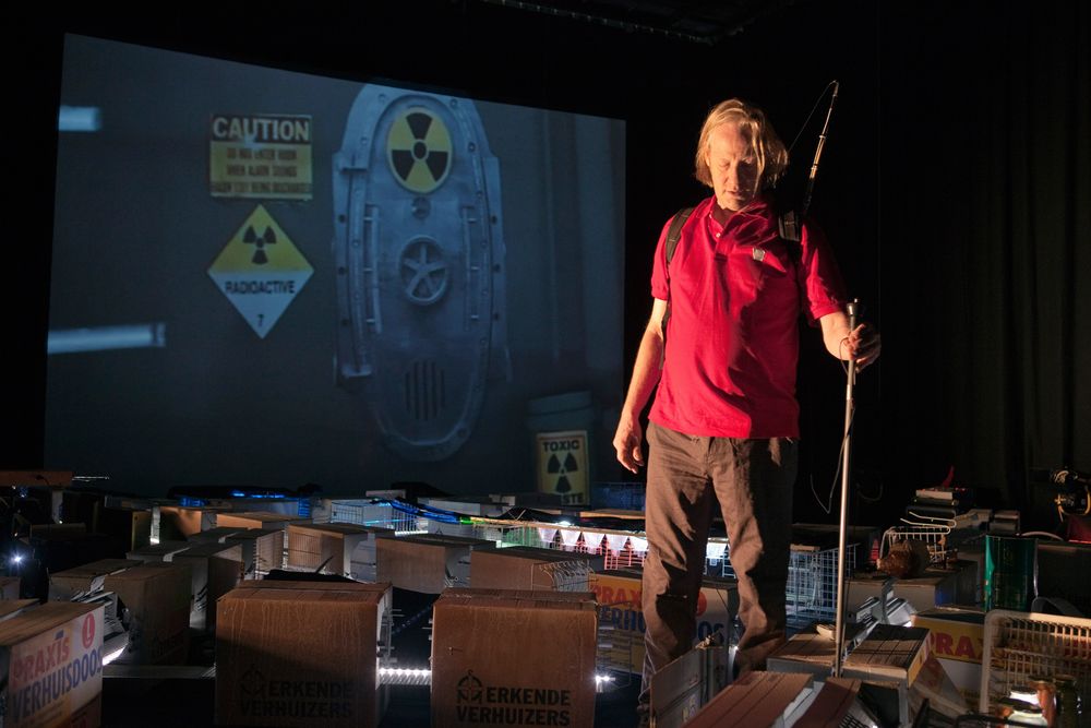 Een speler in een rood poloshirt loopt met een stok over het decor van kratten; op het scherm een vat met het waarschuwingsteken voor radioactiviteit.