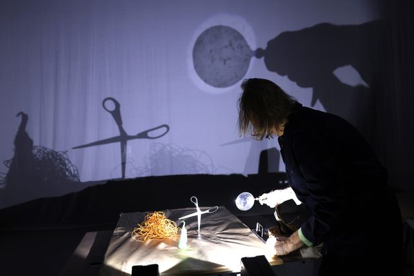 Een speler schikt met een lampje een maantje en een kruisje op de lichttafel; op het doek verschijnen de schaduwen van een schaar en een volle maan.