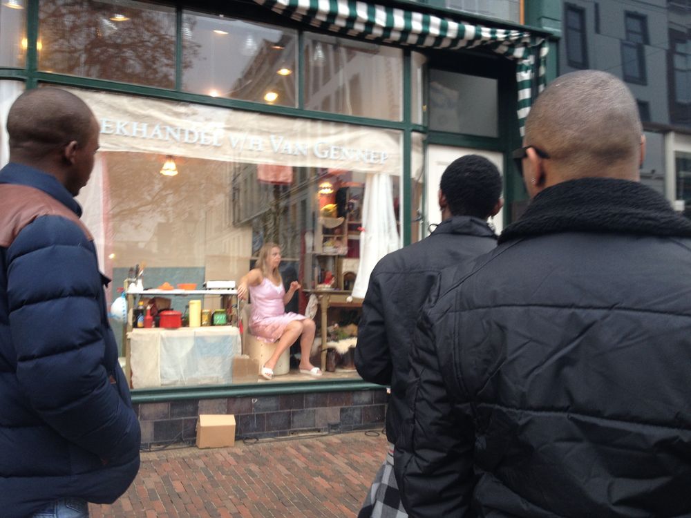 Drie voorbijgangers kijken naar de etalage waarin de vrouw in het roze op haar kruk zit.