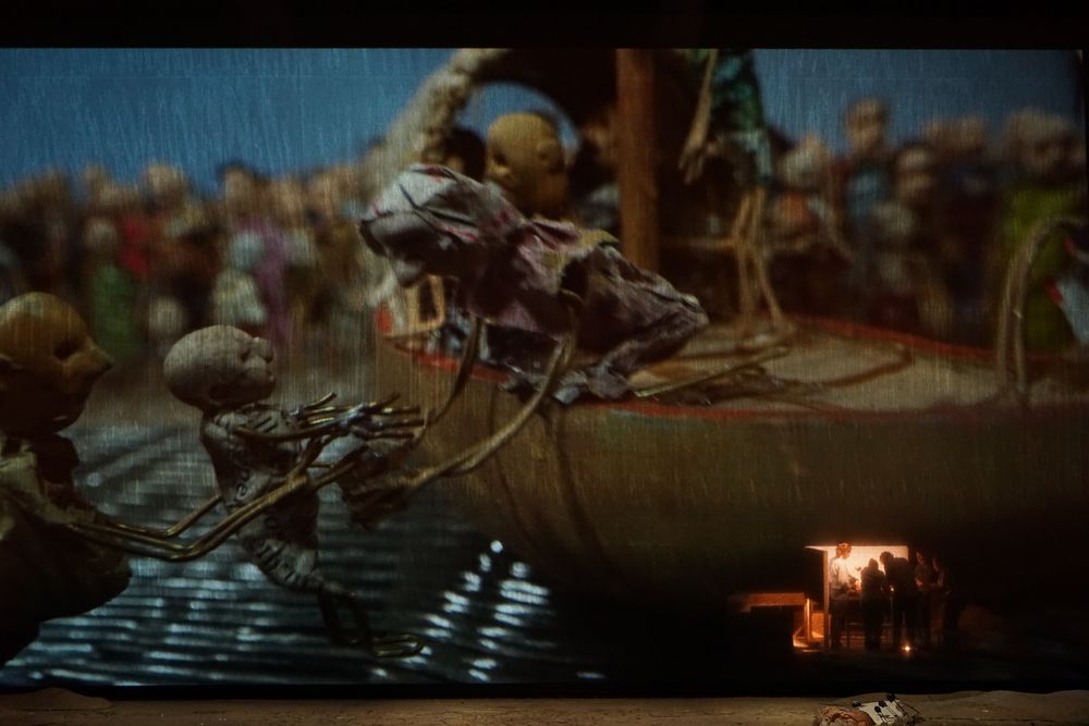 Geprojecteerd beeld: poppen klauteren aan een volle boot in het water, met een menigte figuren op de oever.