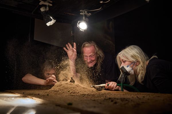 Drie makers boven de zandmaquette: een van hen laat zand door zijn vingers stuiven, rechts houdt een tweede met stofmasker een blaaspijp vast.
