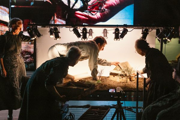 Vier spelers werken onder een rij spots aan een verlicht miniatuurlandschap met een huisje; op het scherm erboven een roodgekleurde close-up.