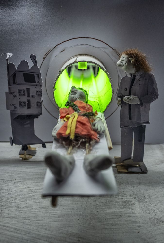Miniatuur-scanner met een groen oplichtende opening; op de schuiftafel ligt een pop, ernaast staat een dokterspop in witte jas en links een bedieningspaneel.