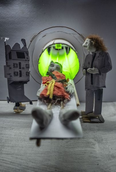 Miniatuur-scanner met een groen oplichtende opening; op de schuiftafel ligt een pop, ernaast staat een dokterspop in witte jas en links een bedieningspaneel.