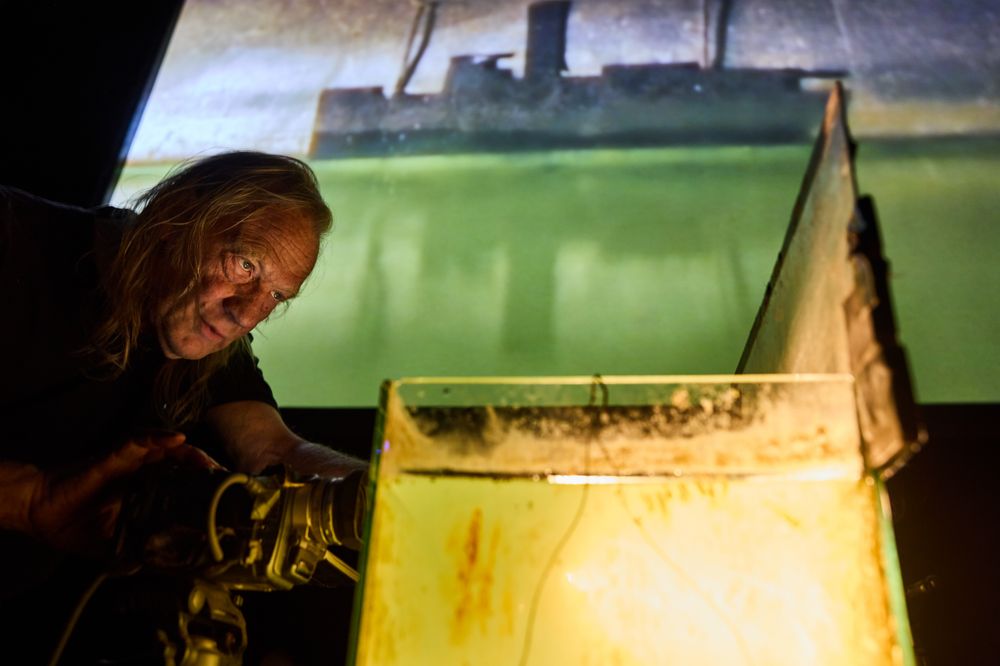 Een maker filmt van dichtbij een waterbak met een scheepje; het beeld ervan staat groot en groengeel op het scherm achter hem.