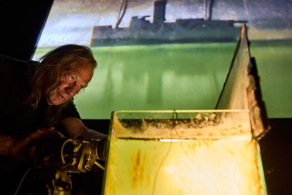 Een maker filmt van dichtbij een waterbak met een scheepje; het beeld ervan staat groot en groengeel op het scherm achter hem.