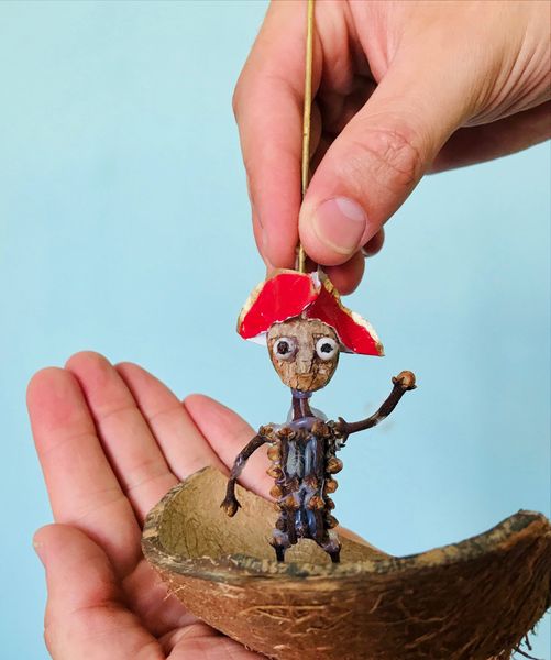 Een hand laat een poppetje met een rode steek zakken in een bootje van een kokosnootschaal dat op een andere hand rust.