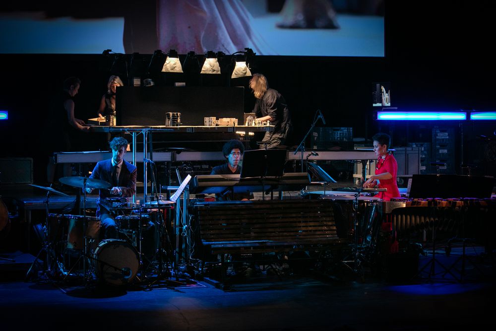 Slagwerkers spelen op drums en marimba onder de verlichte werktafel waar de beelden worden gemaakt.