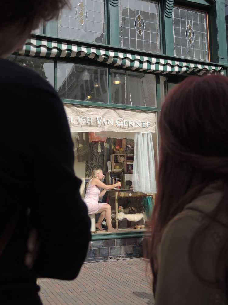 Tussen twee toeschouwers door is de etalage te zien waarin de vrouw in het roze op haar kruk zit.