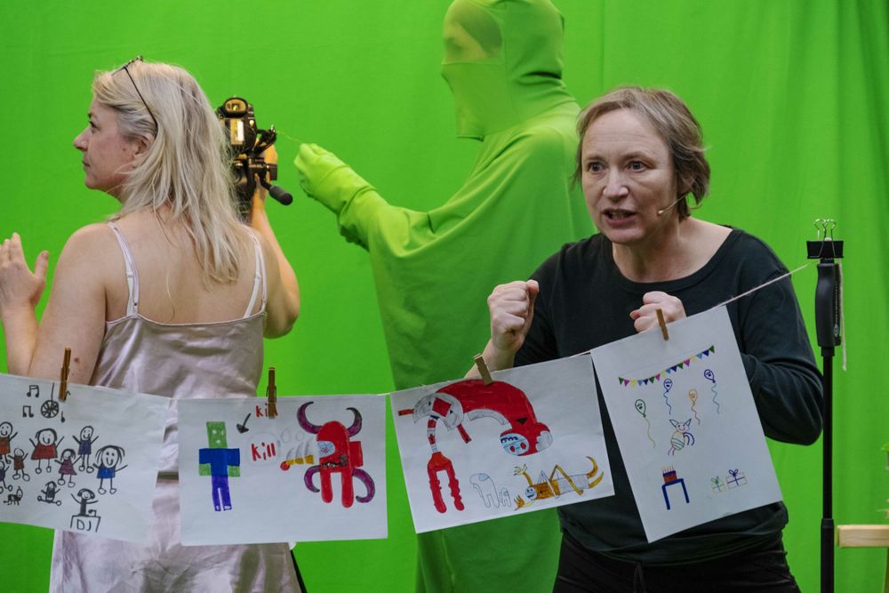 Twee speelsters houden kindertekeningen aan een waslijn omhoog voor het groene doek; achter hen staat iemand in een groen pak.