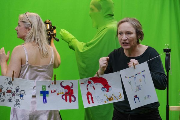 Twee speelsters houden kindertekeningen aan een waslijn omhoog voor het groene doek; achter hen staat iemand in een groen pak.