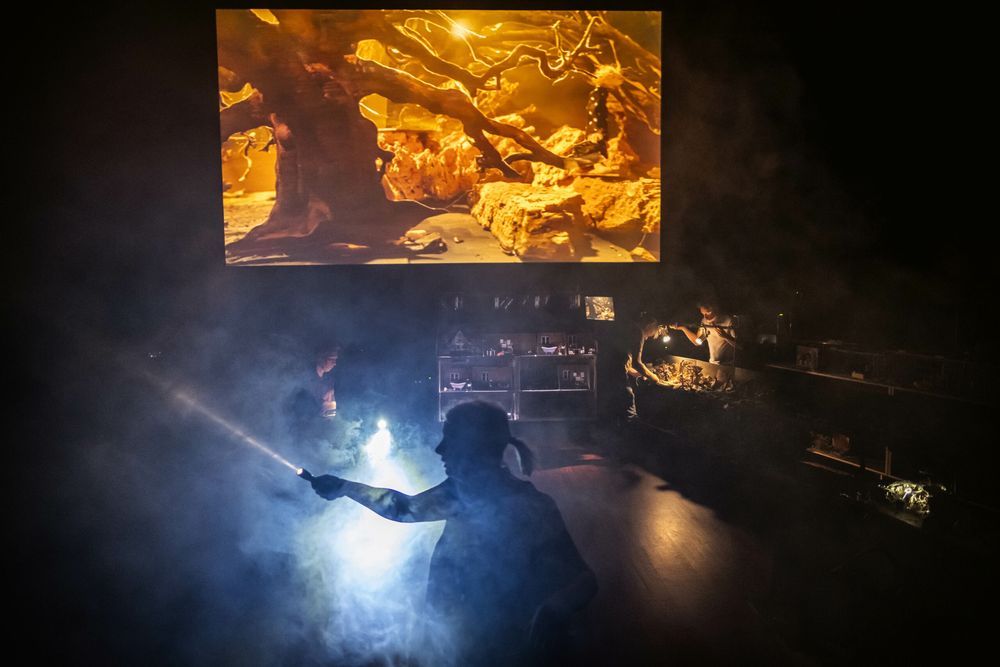 Toneelbeeld: op het grote scherm een goudgeel landschap van wortels en rotsen, daaronder een speler als silhouet met een lamp in de rook, tussen de filmsets.