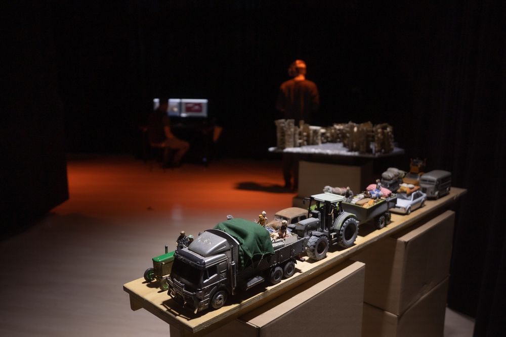Miniatuurvrachtwagens en tractoren vol bagage staan op een sokkel in het donker; achterin zit iemand aan een laptop en staat een tweede in het rode licht.