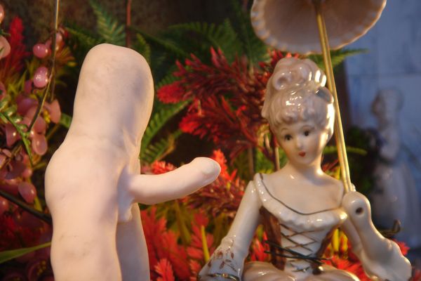 Een wit naakt beeldje steekt zijn hand uit naar een porseleinen dame met een parasol, tussen rode varens.