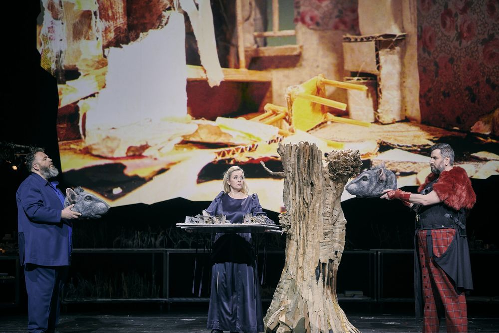 Michael König, Simone Schneider en Goran Jurić houden rattenkoppen in hun handen; in het midden zit een zangeres aan een tafeltje, erachter de projectie van een verwoest interieur.
