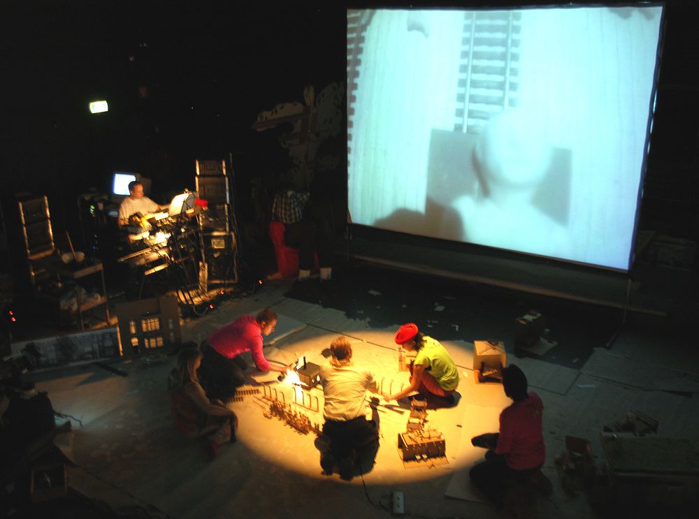 Blik van boven op het toneel: spelers knielen rond een verlicht miniatuurdecor op de witte vloer, links de geluidstafel, rechts het grote projectiescherm.