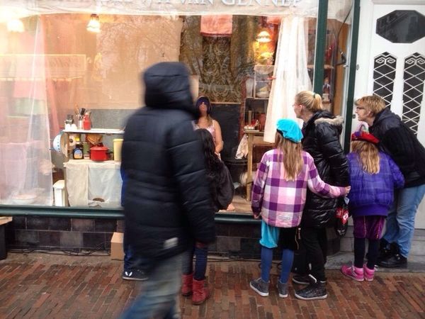 Voorbijgangers en kinderen in winterjassen kijken van de stoep in de etalage waar een vrouw in het roze woont.