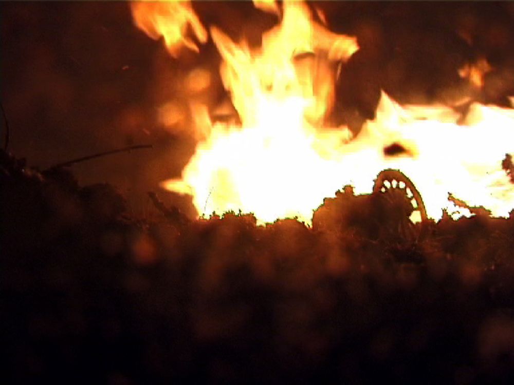Close-up van vlammen boven het miniatuurlandschap; in het tegenlicht tekent zich het wiel van een kanon af.