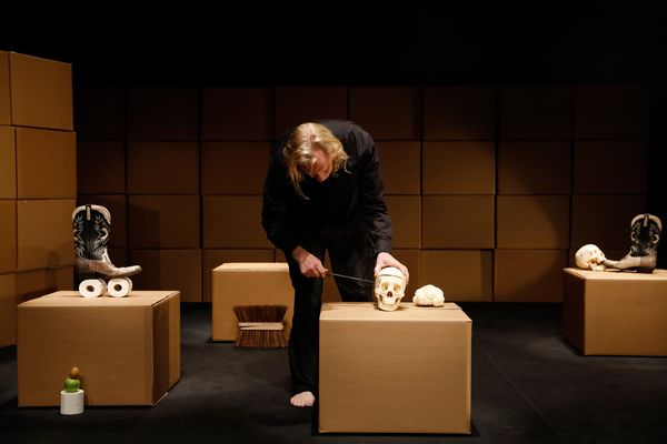 Herman Helle buigt zich met een mes over een kartonnen doos waarop een schedel en een broodje liggen; om hem heen cowboylaarzen, wc-rollen en een luiwagen op andere dozen.