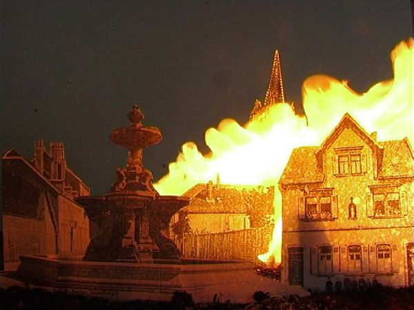 Een miniatuurstad in brand: op het plein staat een fontein, daarachter slaan de vlammen uit de huizen en de kerktoren.