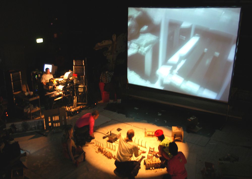 Blik van boven op het toneel: rond het verlichte decor zitten vijf spelers, op het scherm het beeld van omgevallen witte meubels.