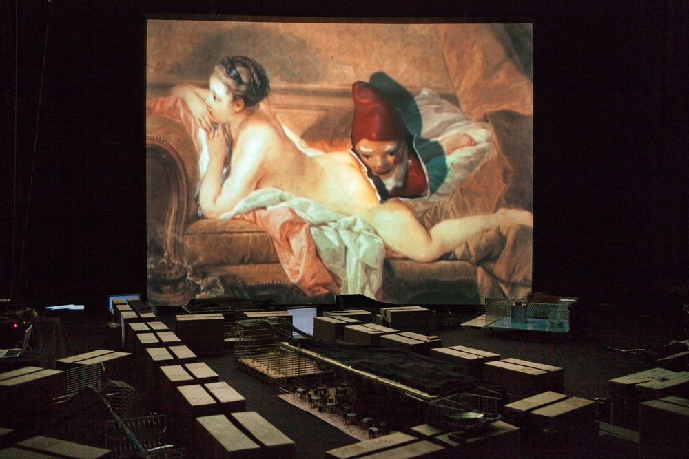 Op het grote scherm hangt een rococoschilderij van een liggende naakte vrouw, daaronder ligt de stad van kratten en dozen in het donker.