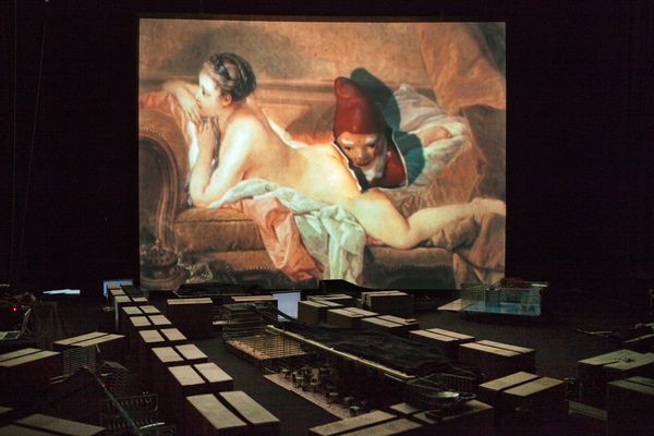 Op het grote scherm hangt een rococoschilderij van een liggende naakte vrouw, daaronder ligt de stad van kratten en dozen in het donker.