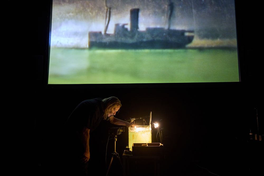 Wijd toneelbeeld: op het scherm een schip in groen water, daaronder buigt een maker zich over de kleine verlichte waterbak.