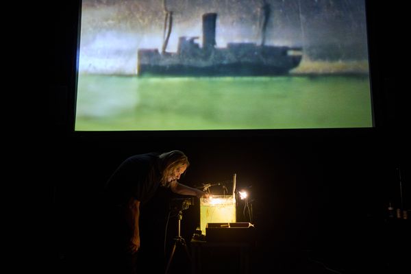 Wijd toneelbeeld: op het scherm een schip in groen water, daaronder buigt een maker zich over de kleine verlichte waterbak.