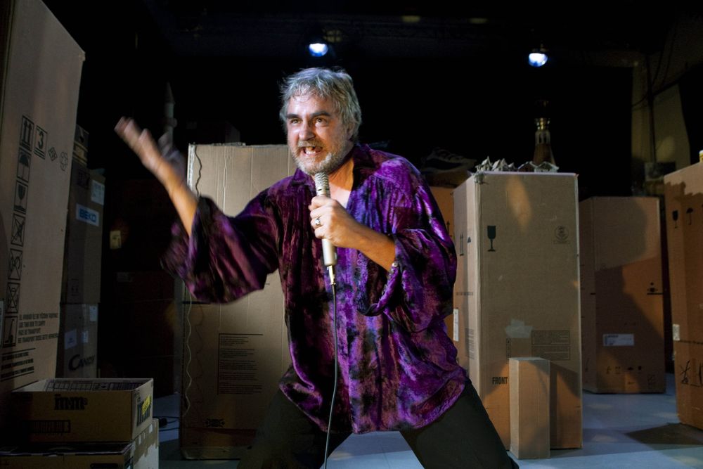 Een man in een paars fluwelen hemd zingt met uitgestoken arm in de microfoon, tussen de kartonnen dozen.