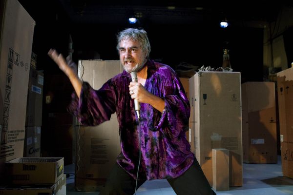 Een man in een paars fluwelen hemd zingt met uitgestoken arm in de microfoon, tussen de kartonnen dozen.
