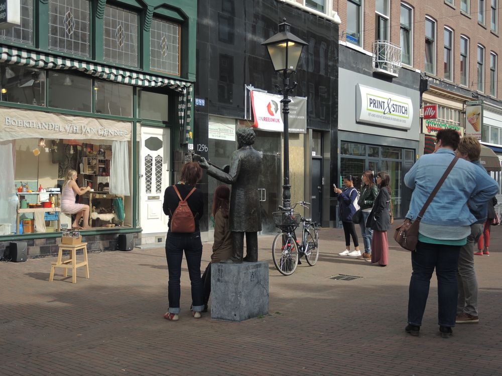 Straatbeeld met links de etalage waarin de vrouw in het roze zit; op het plein een standbeeld en voorbijgangers.