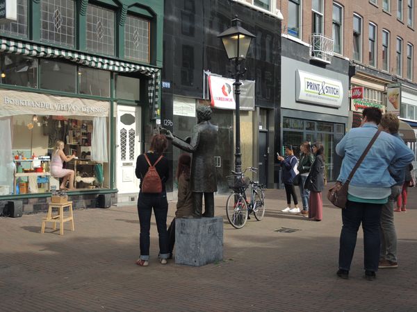 Straatbeeld met links de etalage waarin de vrouw in het roze zit; op het plein een standbeeld en voorbijgangers.